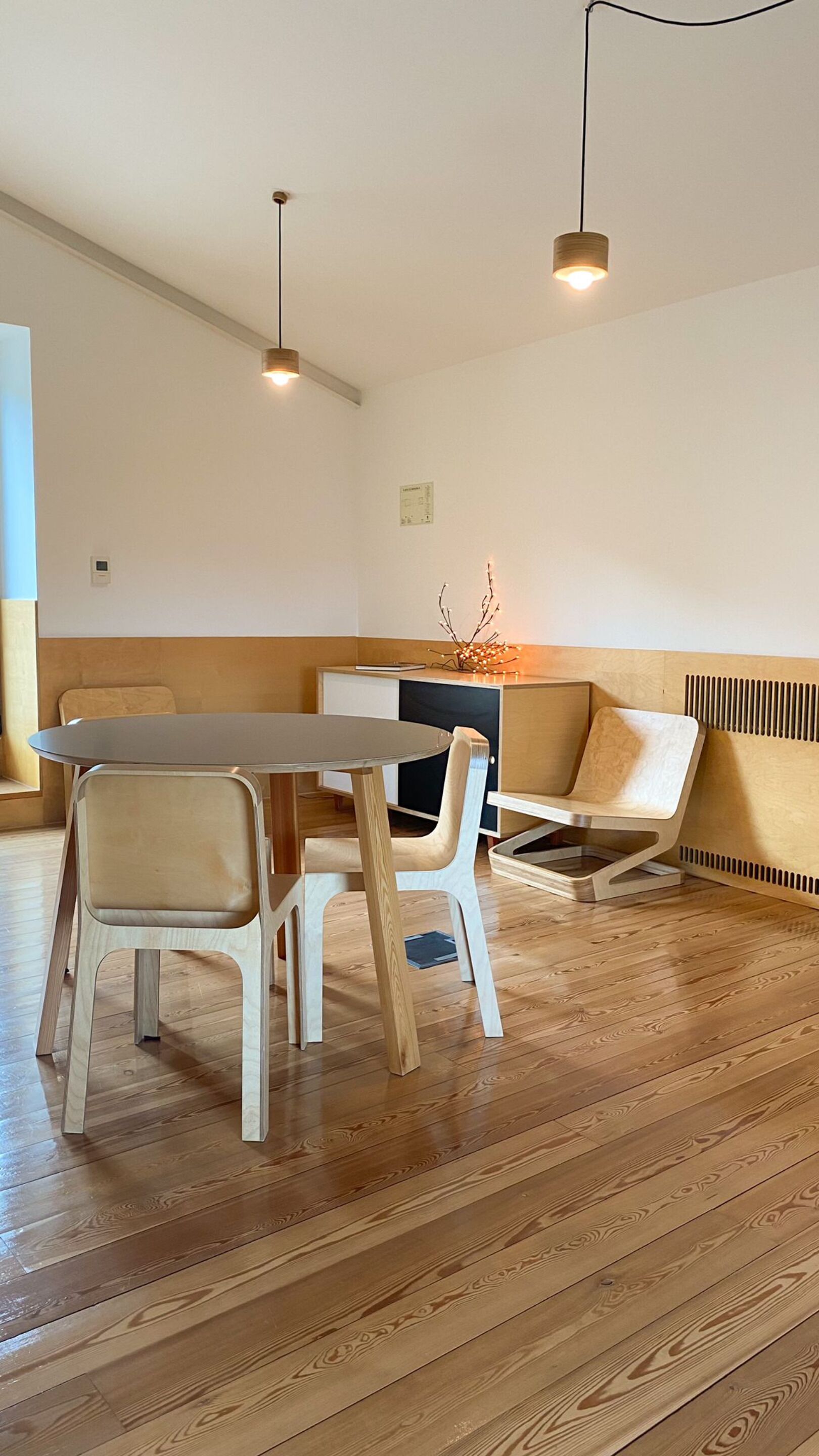Een moderne, minimalistische kamer met houten vloer, een ronde tafel, twee stoelen, een bank en hanglampen.