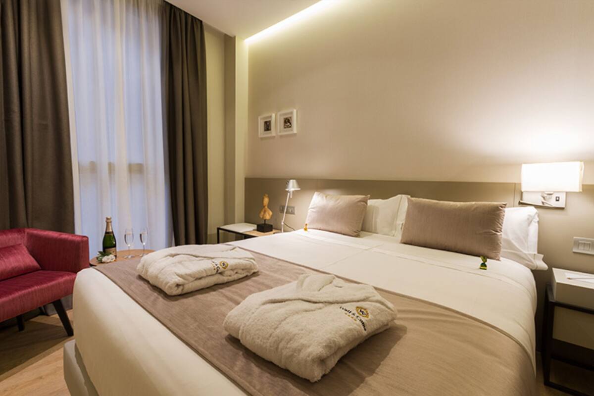 A hotel room with a bed, a red armchair, a nightstand, a lamp, and a window with curtains.