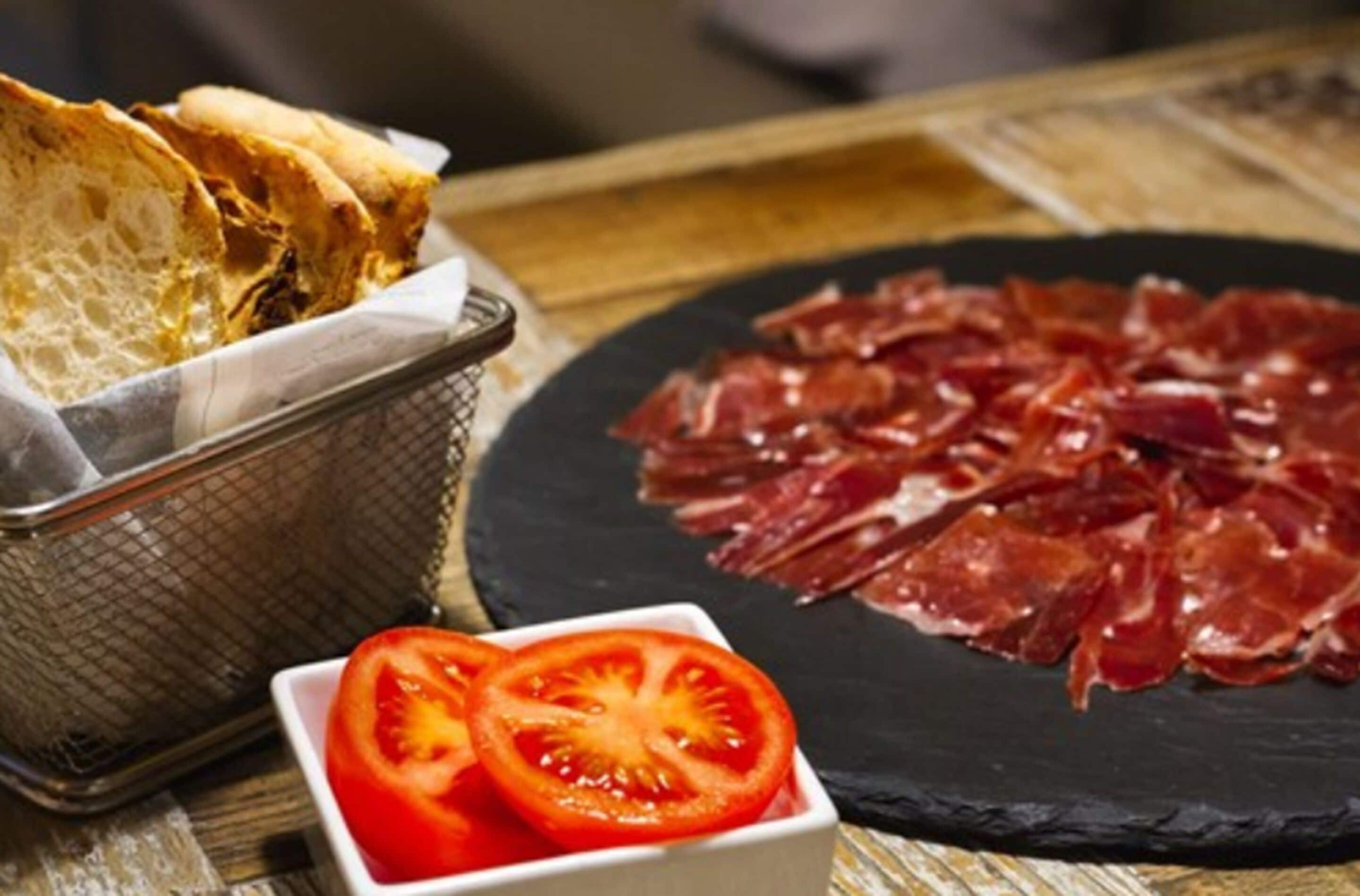 Una tabla de fiambres con carne en rodajas, un bol de rodajas de tomate y una cesta de pan.