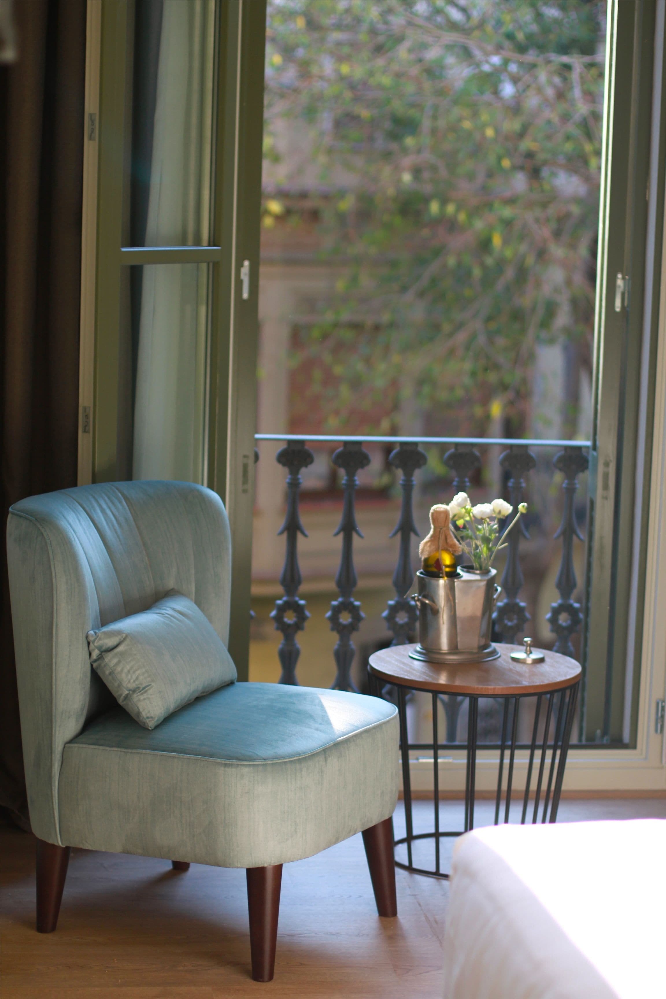 Una habitación con un sillón azul, una mesita redonda con un jarrón de flores y vistas a un balcón con barandilla de metal.