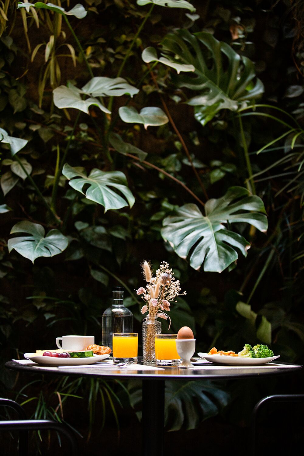 A table set with breakfast items, a bottle, and a vase with dried flowers, surrounded by lush greenery.