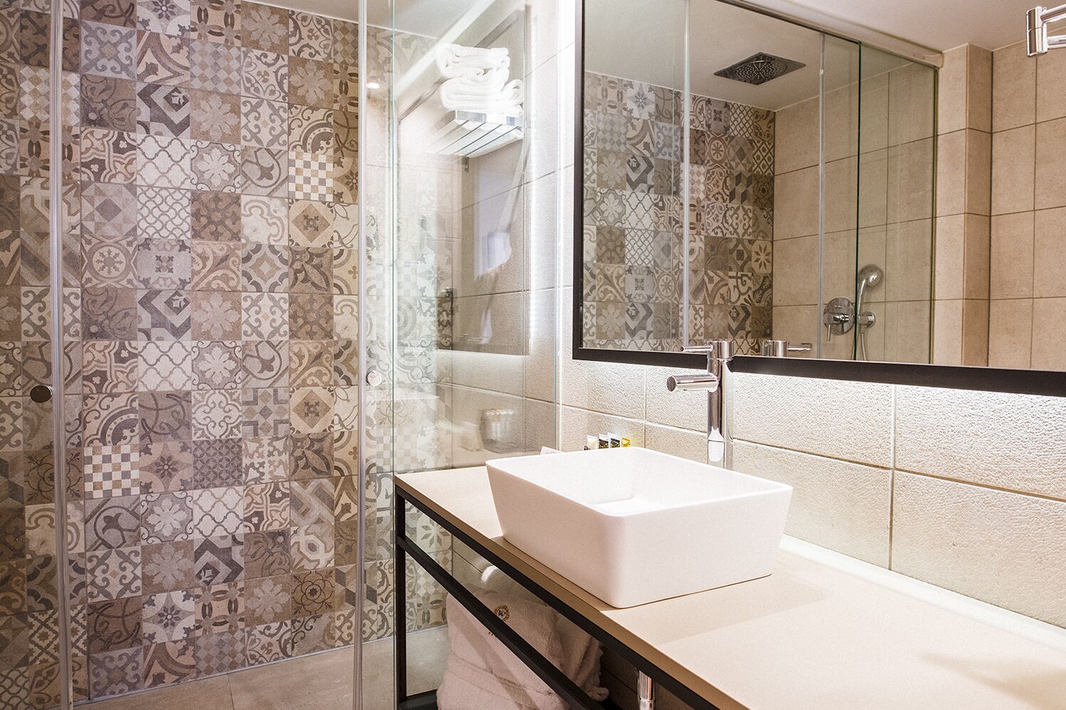 A modern bathroom with patterned tiles, a glass shower enclosure, a white sink, and a large mirror.