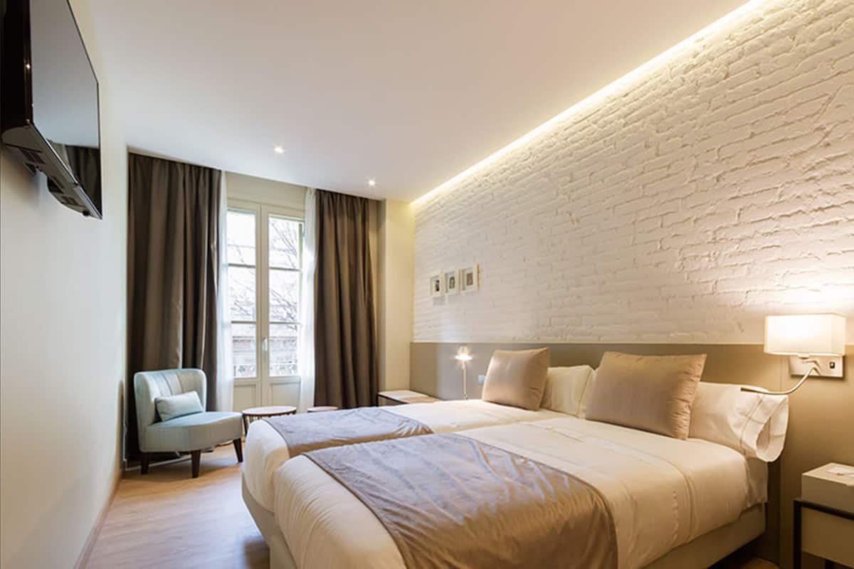 A hotel room with a brick wall, a flat-screen TV, a bed with two pillows, a chair, and a bedside table.