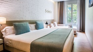 Hotellihuone, jossa on suuri sänky, pienempi sänky, yöpöytä, lamppu, ikkuna, jossa on verhot, ja näkymä vihreälle alueelle.