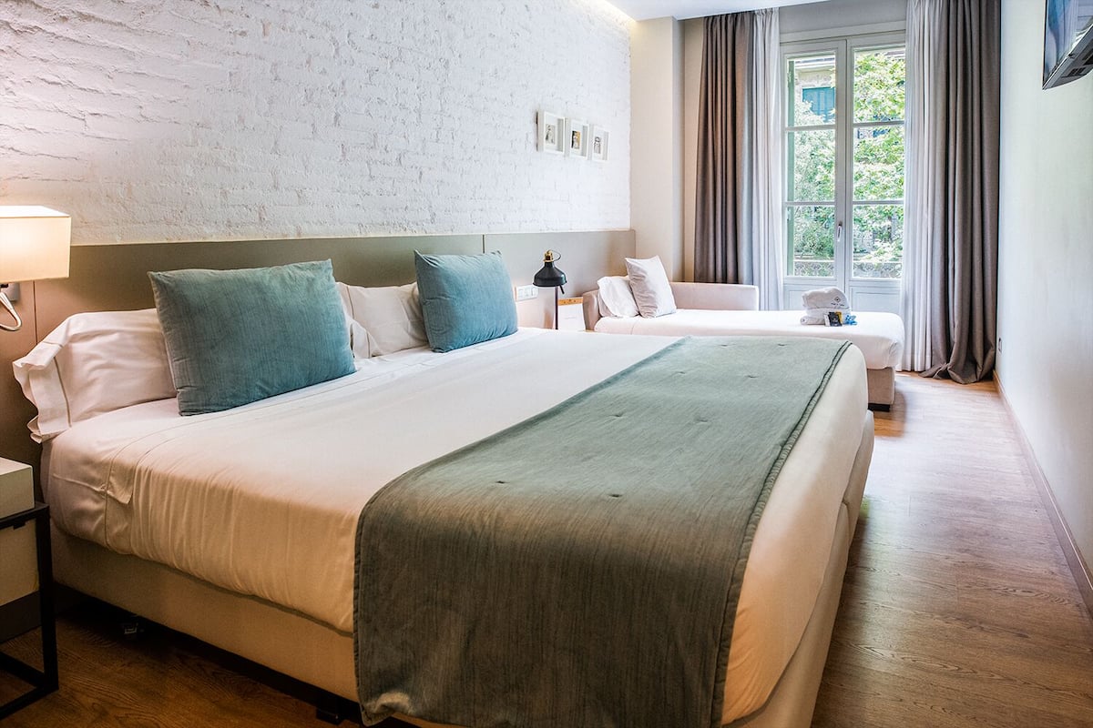 A hotel room with a large bed, a smaller bed, a nightstand, a lamp, a window with curtains, and a view of greenery outside.