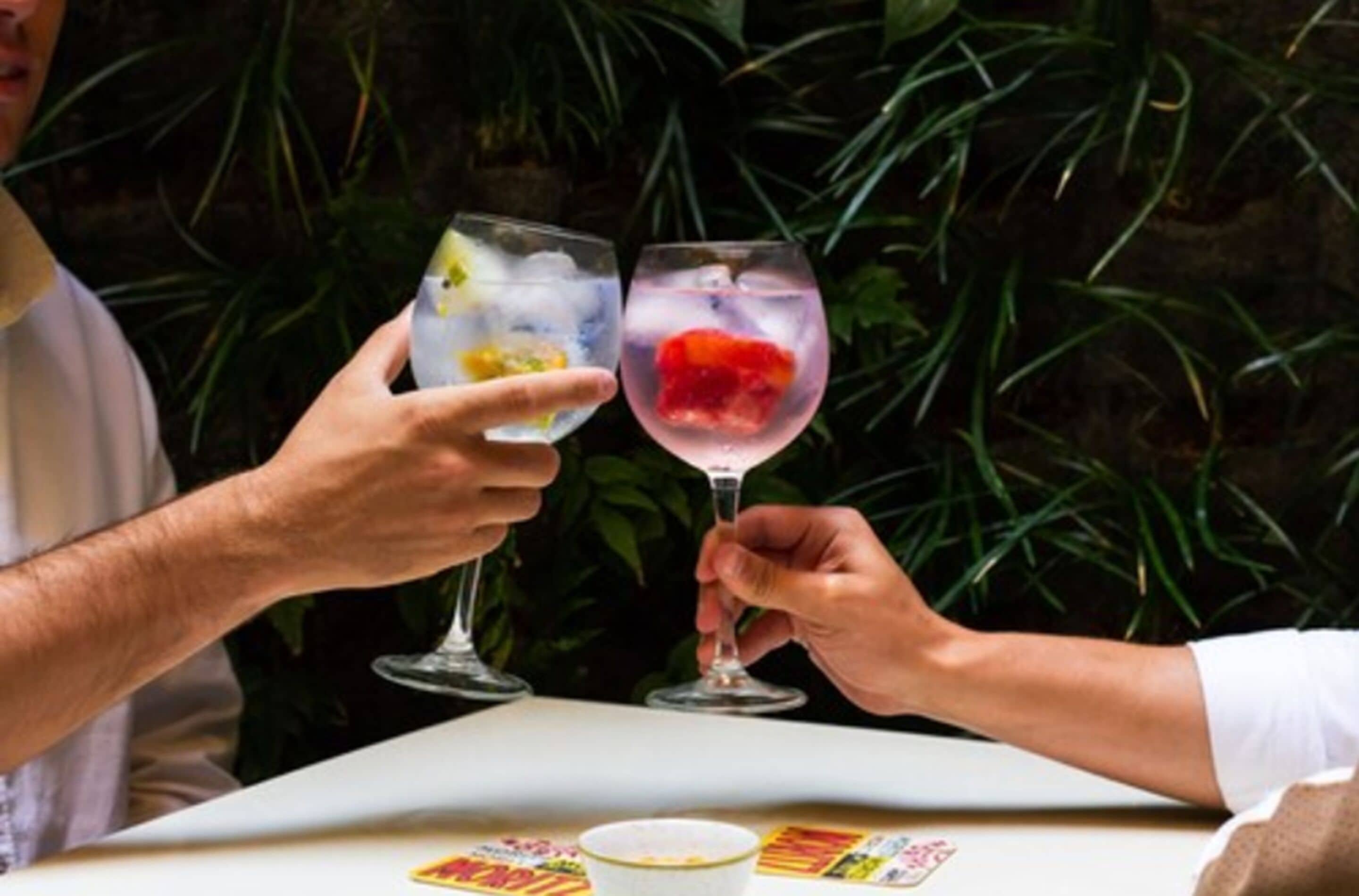 Dos personas sosteniendo vasos con bebidas heladas y fruta, sentadas en una mesa con mantel blanco.