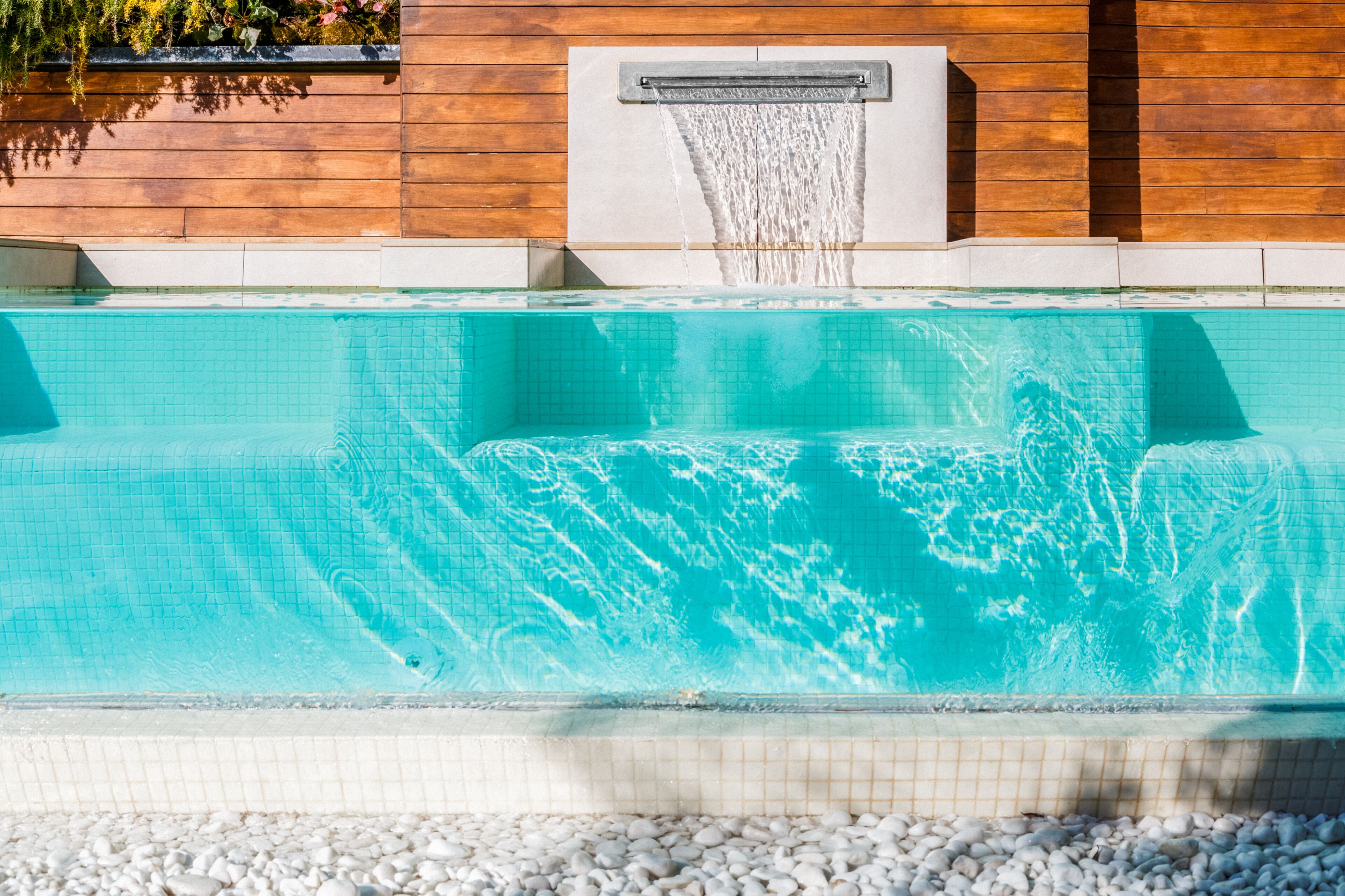 A swimming pool with a waterfall feature, surrounded by a pebble border and wooden walls.