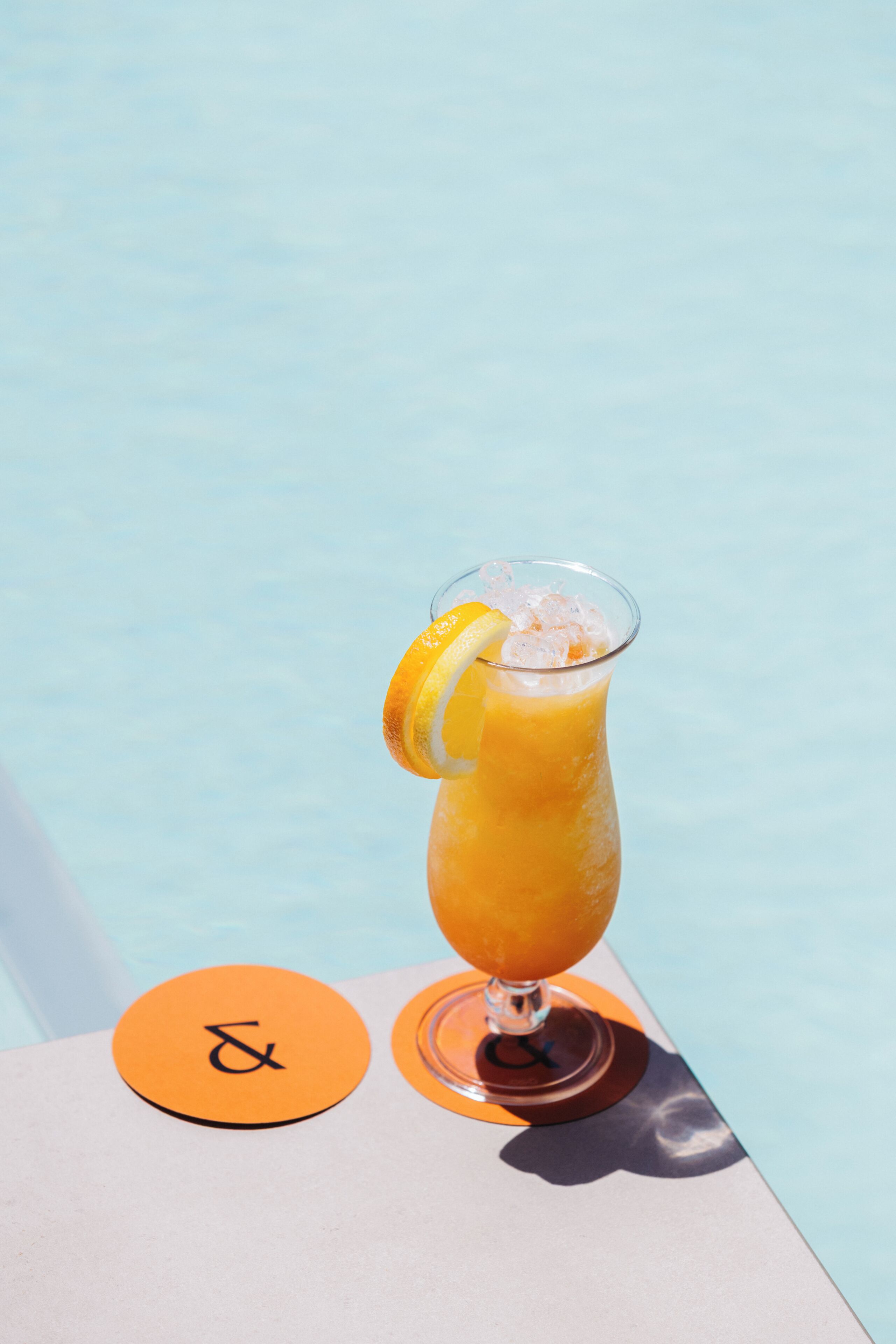 A glass of orange juice with a lemon slice on a table by a pool.