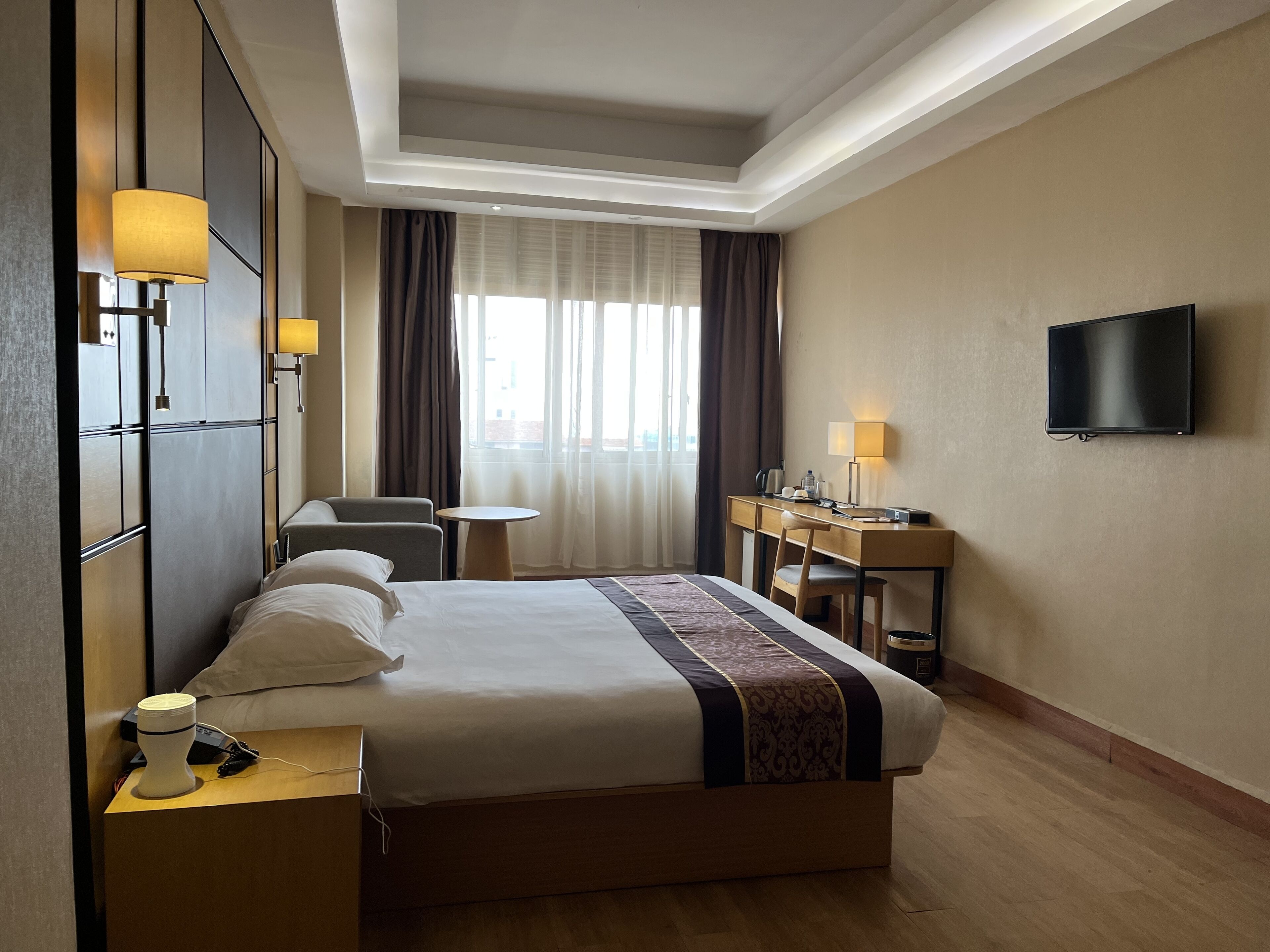 Una habitación de hotel con cama, mesita de noche, lámpara, escritorio, silla, televisor y cortinas.