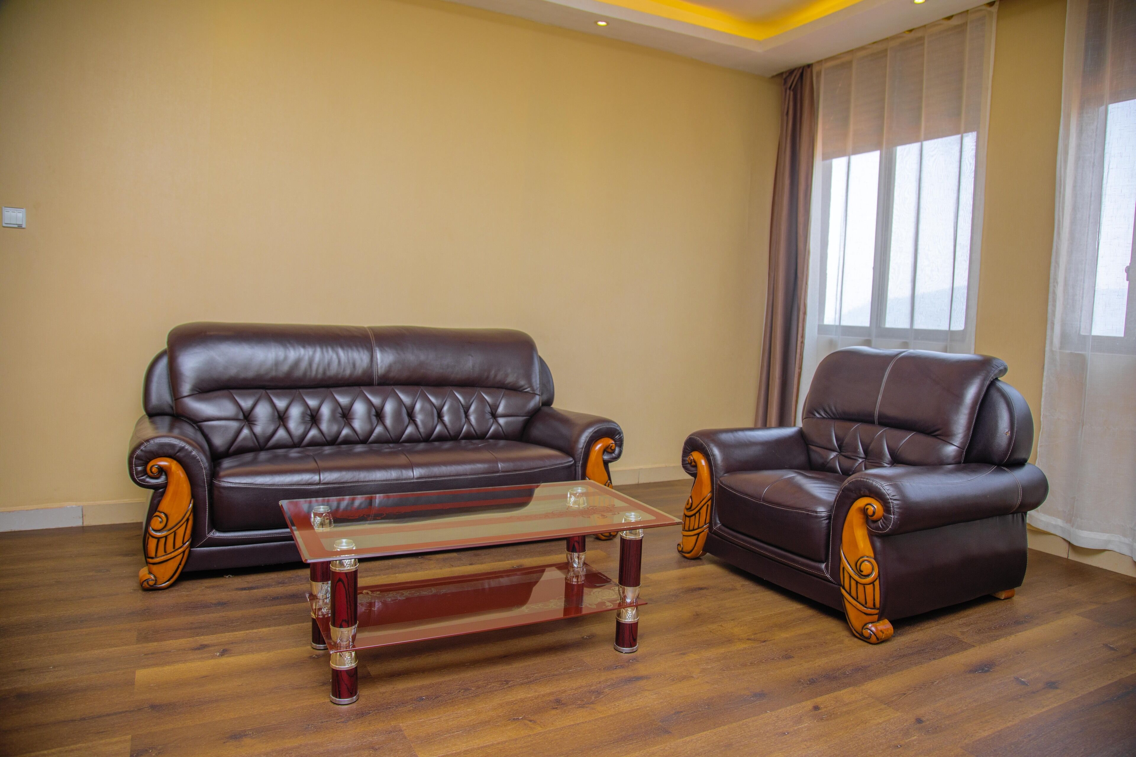 A living room with a brown leather sofa and armchair, a glass coffee table, wooden flooring, and a large window with curtains.