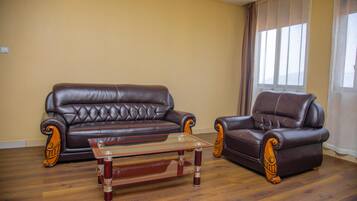 A living room with a brown leather sofa and armchair, a glass coffee table, wooden flooring, and a large window with curtains.