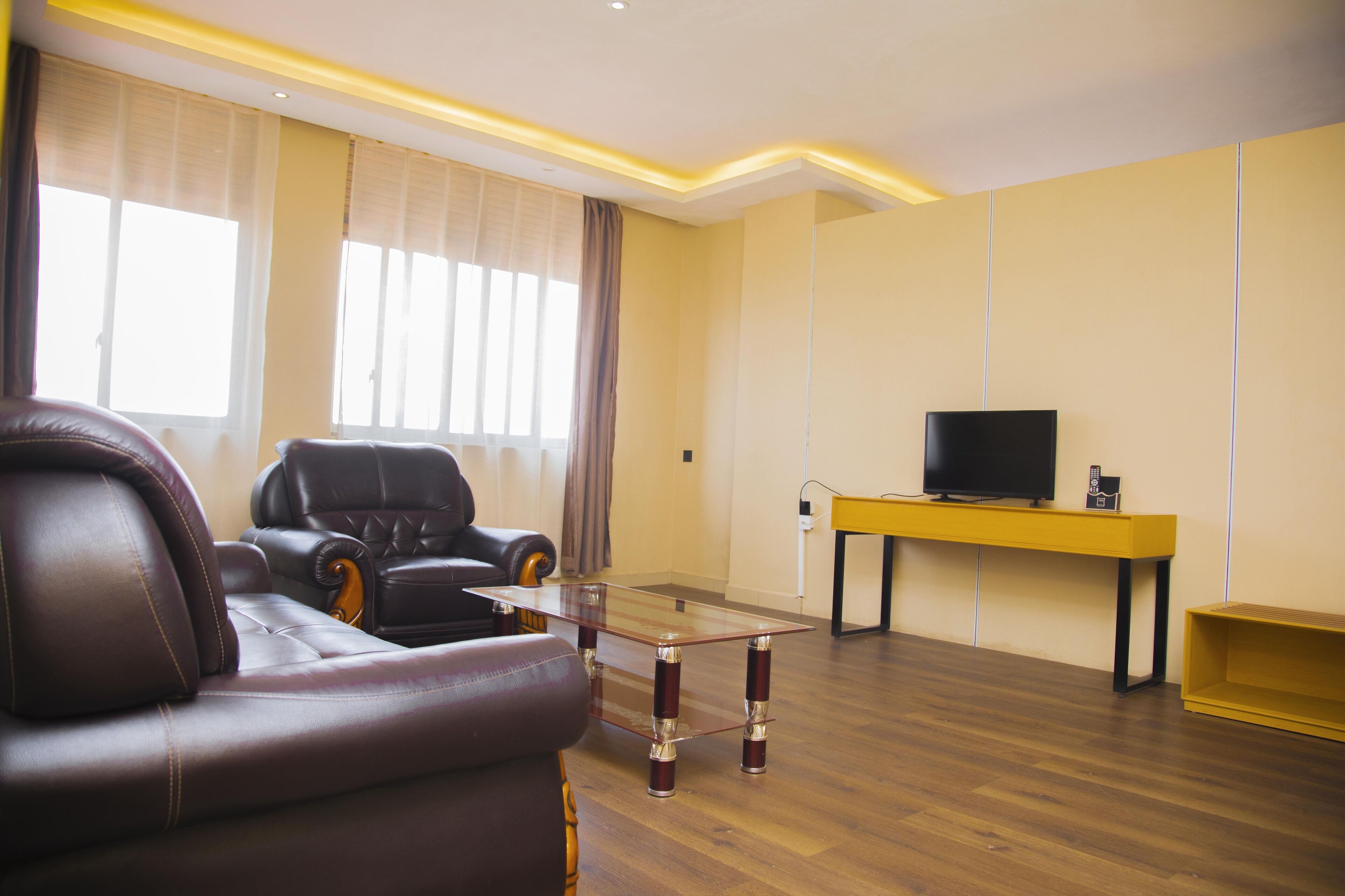 Una sala de estar moderna con sofás de cuero, una mesa de centro de vidrio, un televisor de pantalla plana sobre un mueble amarillo y ventanas amplias con cortinas de tul.