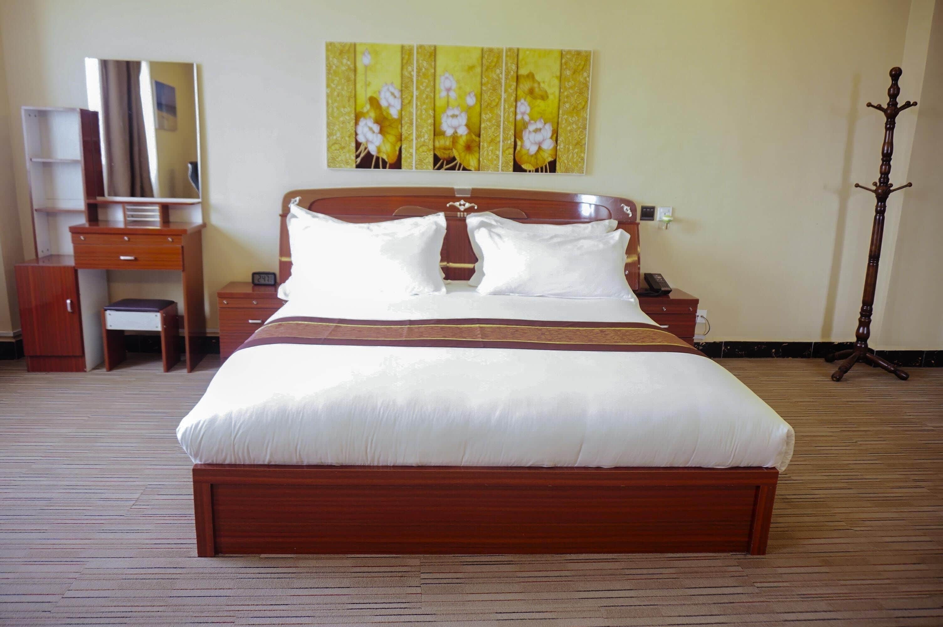 A hotel room with a large bed, wooden headboard, a bedside table, a nightstand, a mirror, a wardrobe, and a painting on the wall.