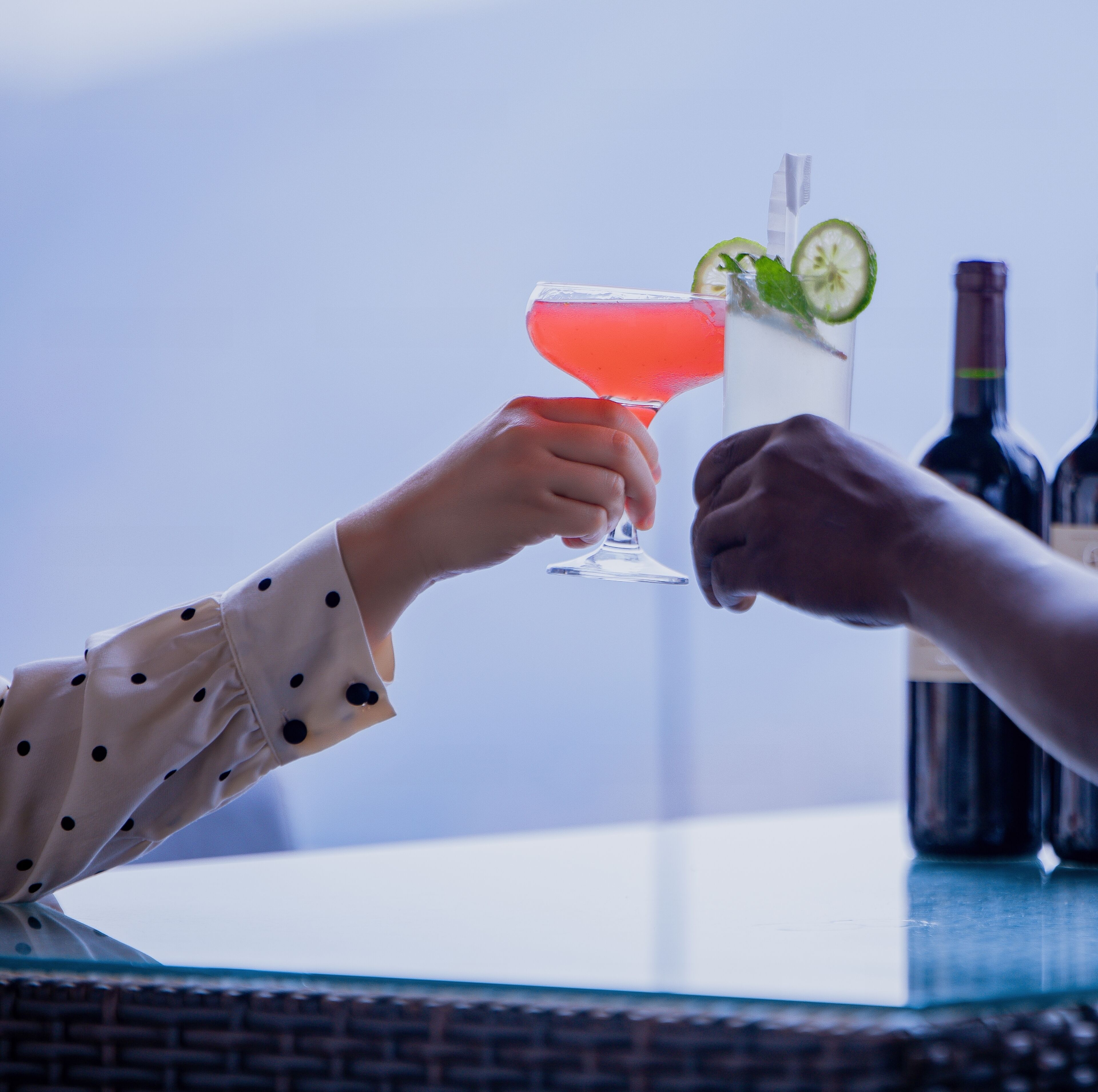 Two hands holding cocktail glasses with a lime garnish, a bottle of wine, and a glass of water on a table.