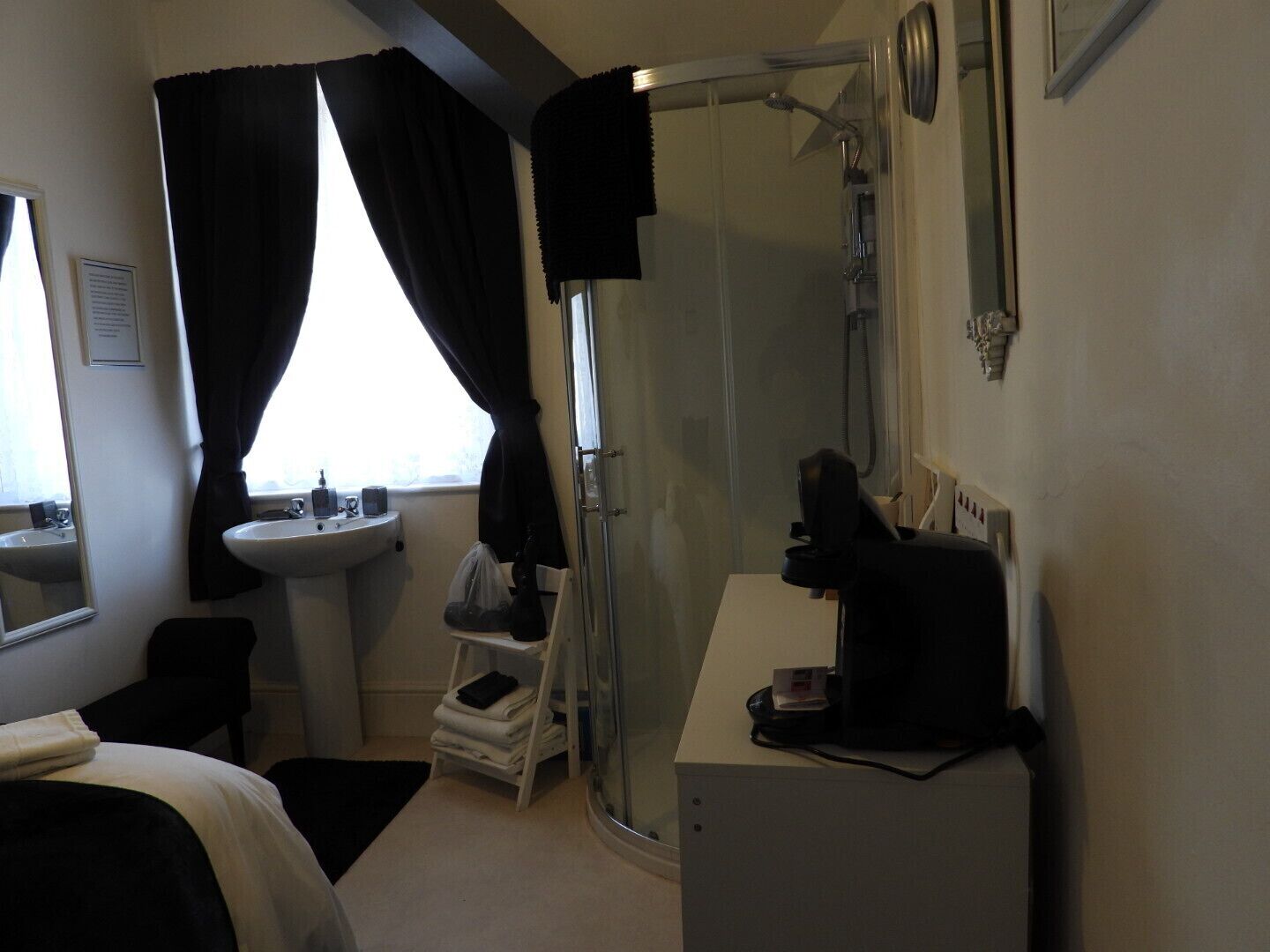 A hotel room with a bed, a bedside table, a shower, a sink, and a window with curtains.