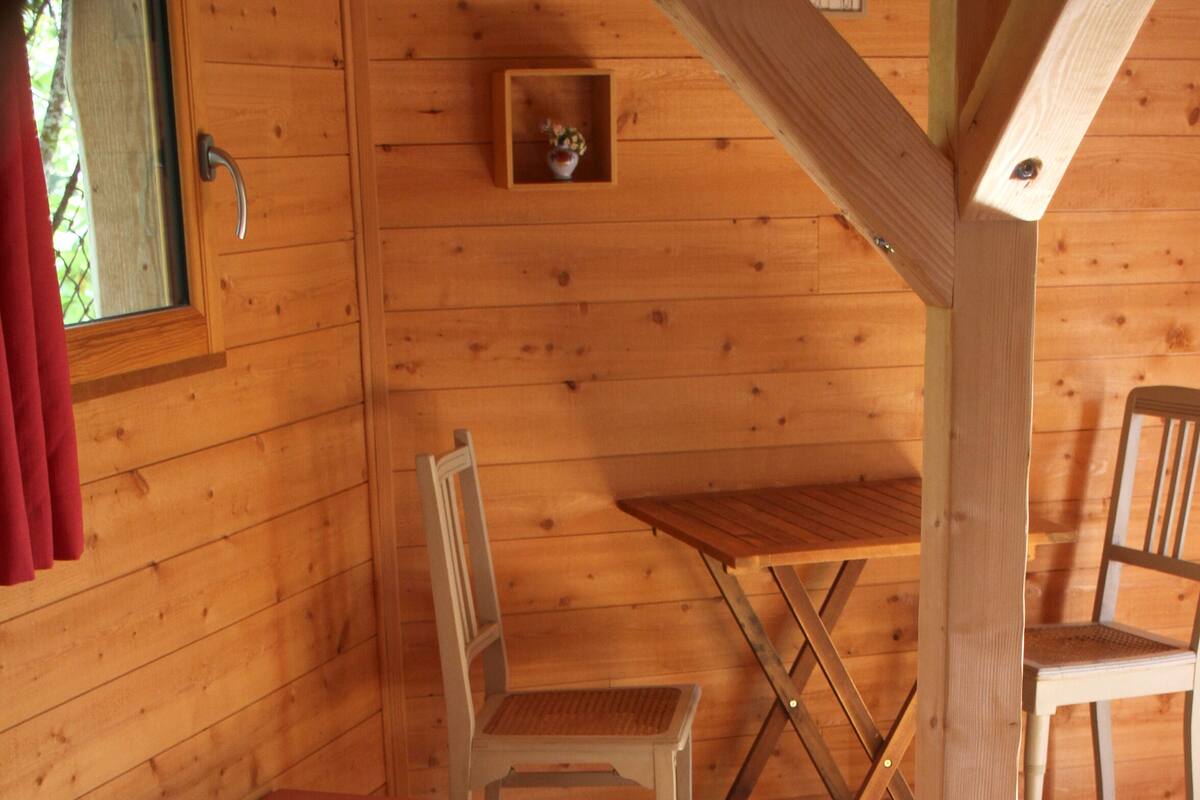 A wooden cabin interior with a bed, a small table, two chairs, and a wooden bench.