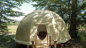 A dome-shaped tent with a wooden walkway leading to the entrance, surrounded by trees and a picnic table in the background. - Ô bois dormant (Lempzours)