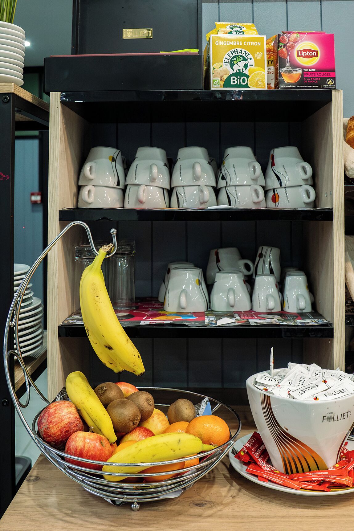 Un coin cuisine avec un panier de fruits contenant des bananes, des pommes et des oranges, une bouilloire et une boîte de thé Lipton.