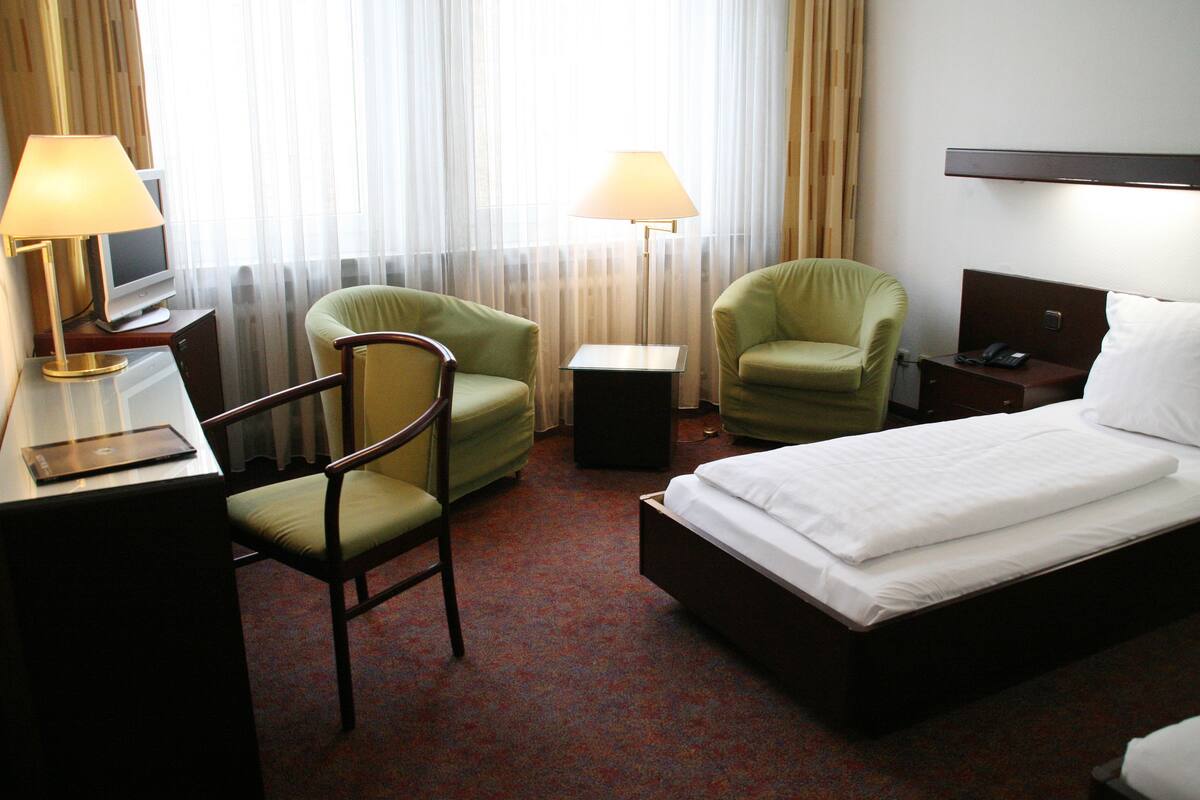 A hotel room with a bed, two chairs, a desk with a lamp, a TV, and a bedside table with a phone and a lamp.