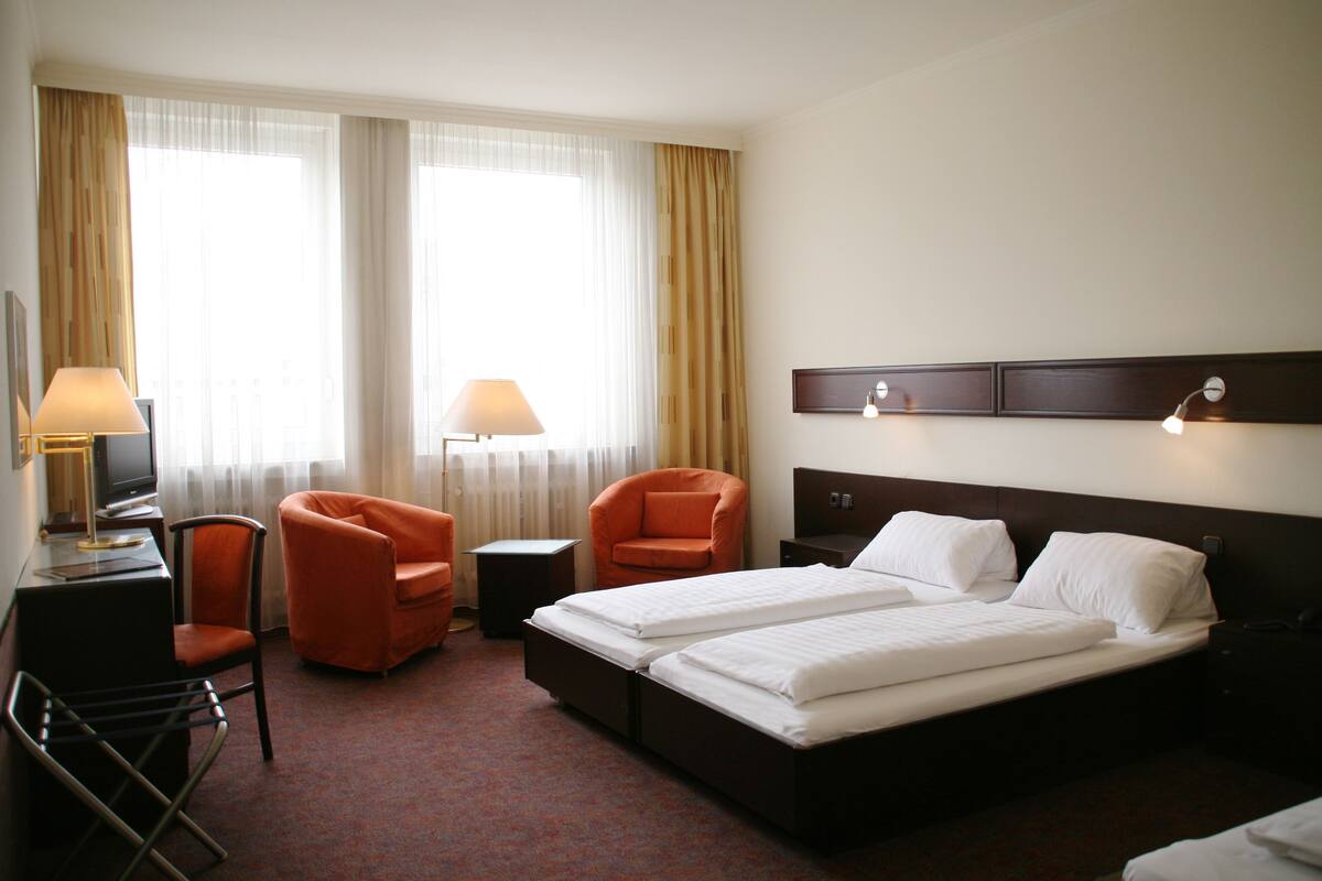A hotel room with two beds, a desk with a lamp, an orange armchair, and a TV.