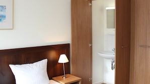 Hotellihuone, jossa on sänky, yöpöytälamppu, puinen vaatekaappi ja kylpyhuone, joka näkyy avoimesta ovesta.