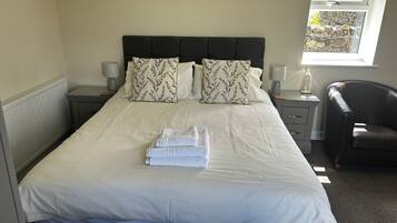 A neatly made bed with a headboard, two bedside tables with lamps, a chair, and a window with a view of a stone wall.