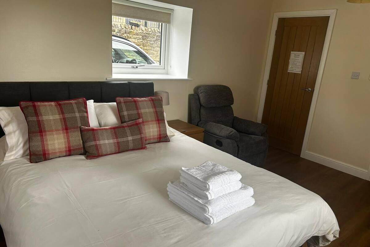 A neatly made bed with plaid pillows, a stack of white towels, a massage chair, and a window with a view of a building outside.
