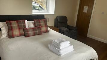 A neatly made bed with plaid pillows, a stack of white towels, a massage chair, and a window with a view of a building outside.