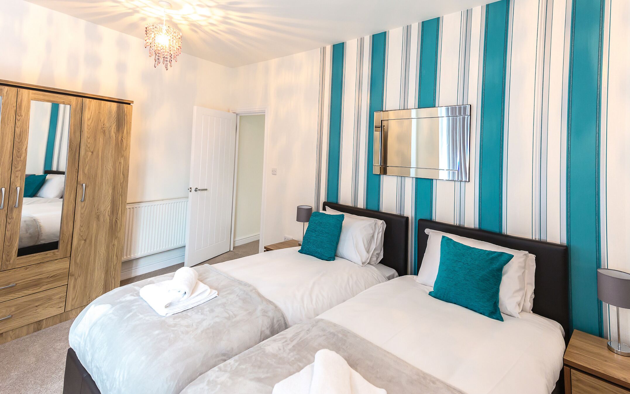 A hotel room with two beds, a wooden wardrobe, a mirror, and a striped wall.