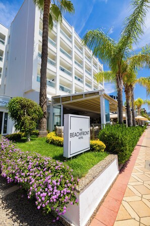 Ein Hotel mit einem Schild, auf dem 'The Beachfront Hotel' steht, umgeben von Palmen und einem gepflegten Garten mit violetten Blumen.