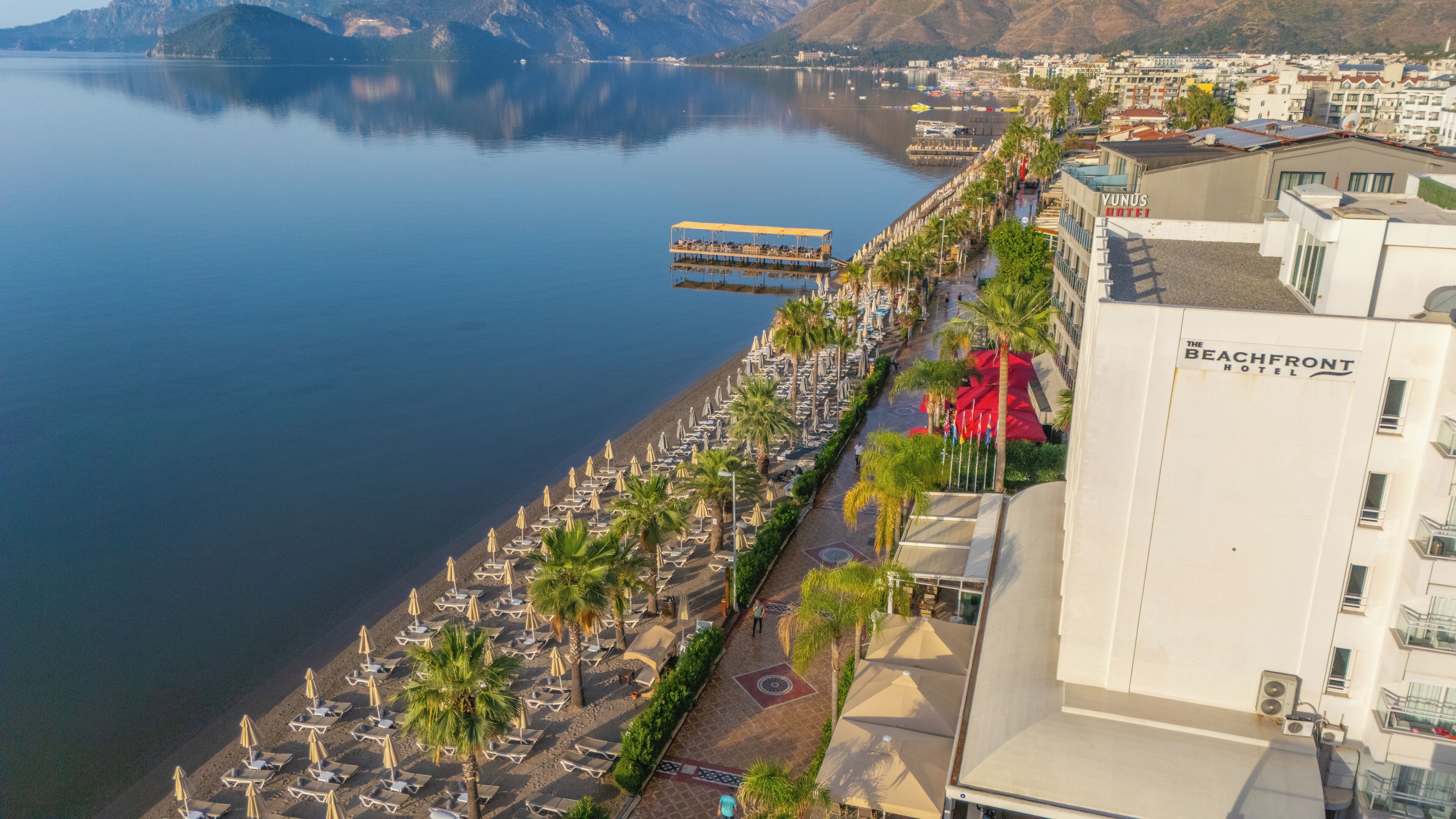 Et strandhotel med række af liggestole og parasoller langs stranden, en mole der stikker ud i vandet, og fri udsigt til de omkringliggende bjerge.