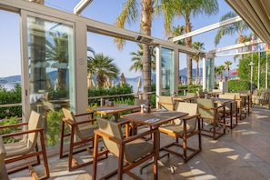 Un restaurante con terraza, grandes ventanales y palmeras.