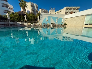 A clear swimming pool with lounge chairs and umbrellas, surrounded by buildings with balconies and a clear sky.
