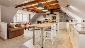 A spacious loft with a high ceiling, exposed wooden beams, a central dining area with white chairs, a kitchen with white cabinets, and a large window providing natural light. - The Tower Gastro Pub & Apartments (Crieff)