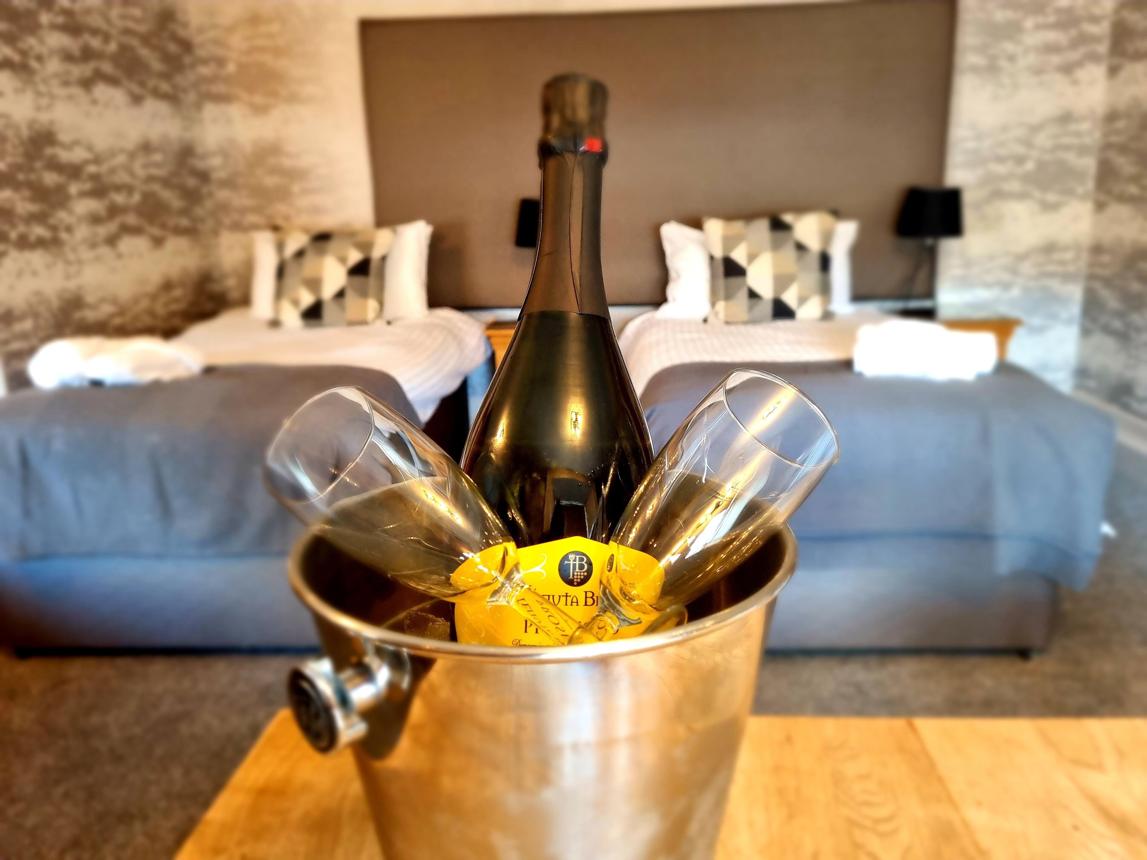 Eine Flasche Champagner in einem Metallkübel mit Gläsern, platziert auf einem Holztisch in einem Hotelzimmer. Im Hintergrund sind ein Bett und ein Nachttisch zu sehen.