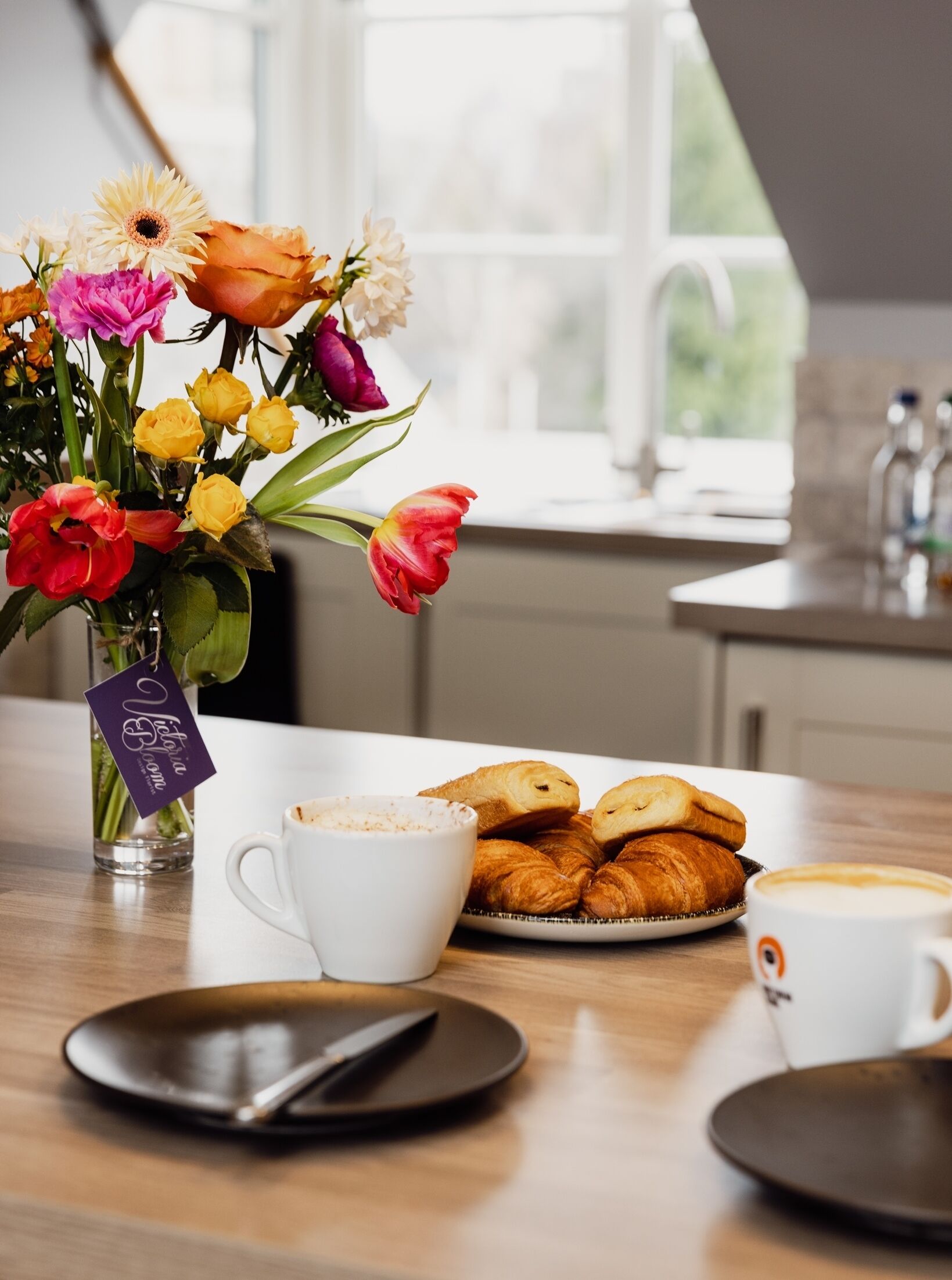 A breakfast setting with a vase of flowers, croissants, and coffee cups on a wooden table.