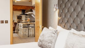 A hotel room with a bed, a tufted headboard, a wooden door leading to a dining area with a table and chairs, and a kitchen in the background. - The Tower Gastro Pub & Apartments (Crieff)