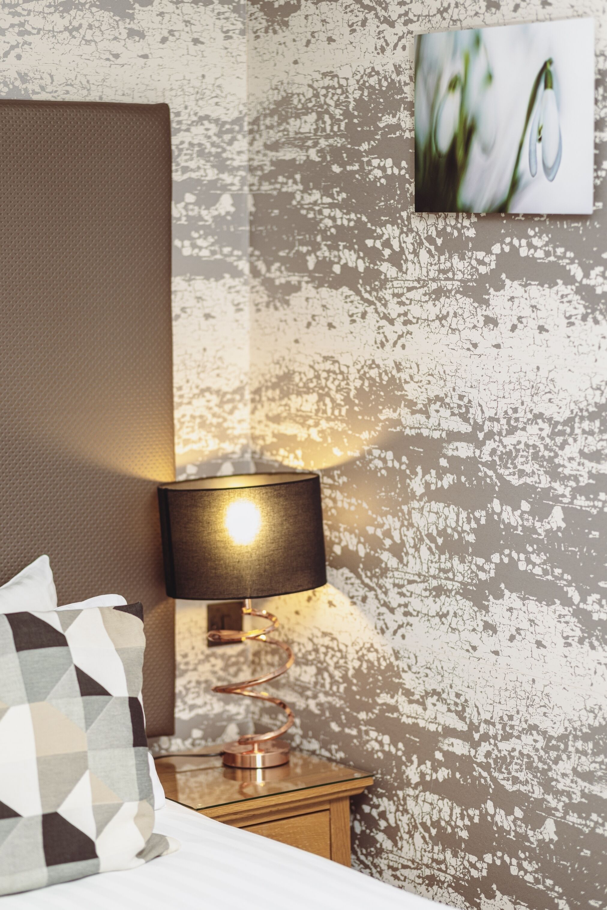 Una cama bien tendida con un cojín estampado con motivos geométricos, una lámpara de pared y un muro texturizado con un cuadro enmarcado.