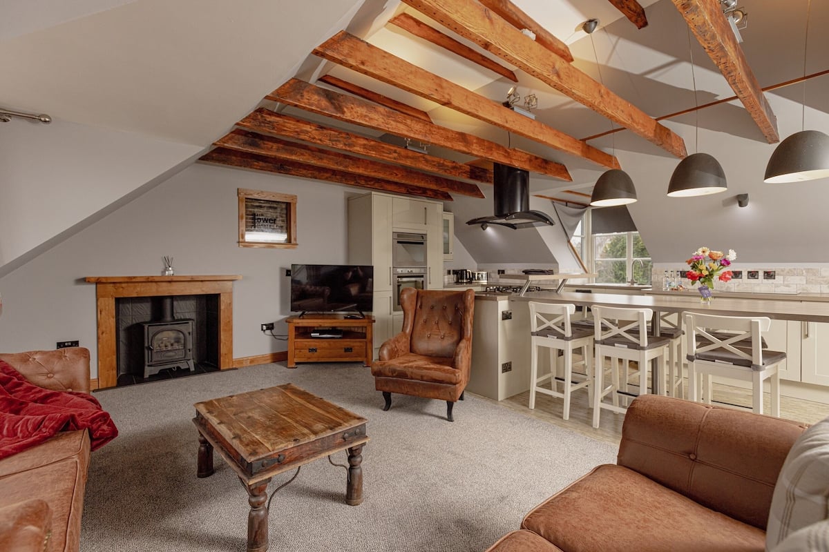A cozy loft-style living area with exposed wooden beams, a fireplace, a kitchen with bar stools, and a comfortable seating area.