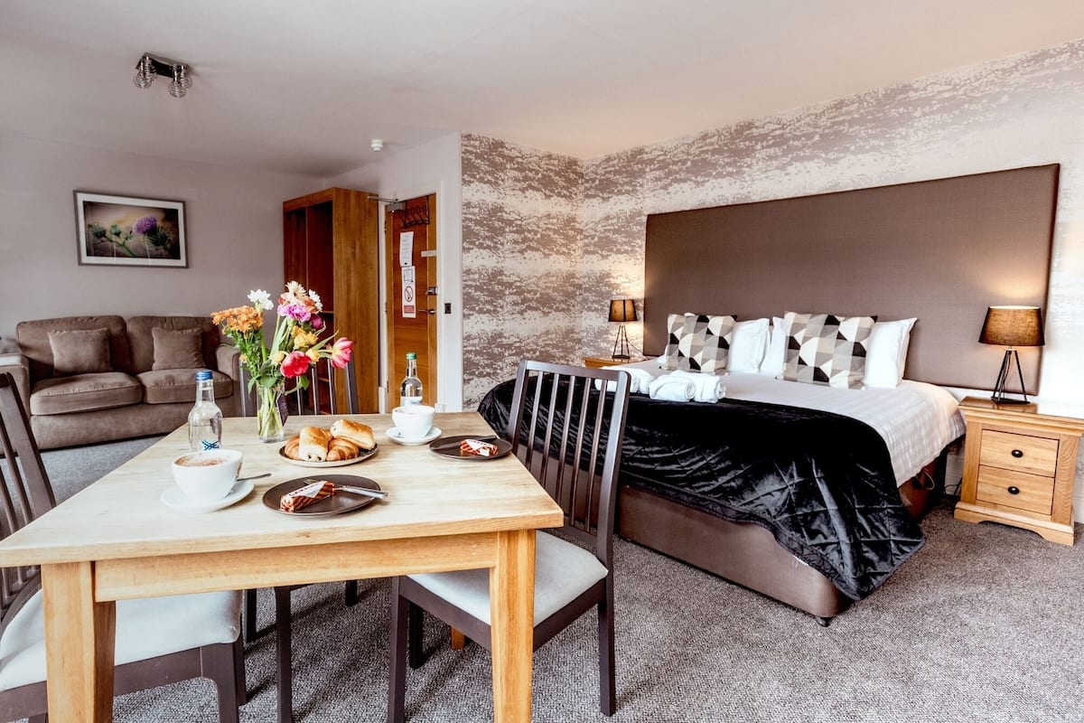 A hotel room with a bed, bedside table, lamp, and a dining area with a table, chairs, and a vase of flowers.