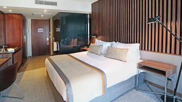 A modern hotel room with a large bed, a desk, a chair, a bedside table, and a lamp.