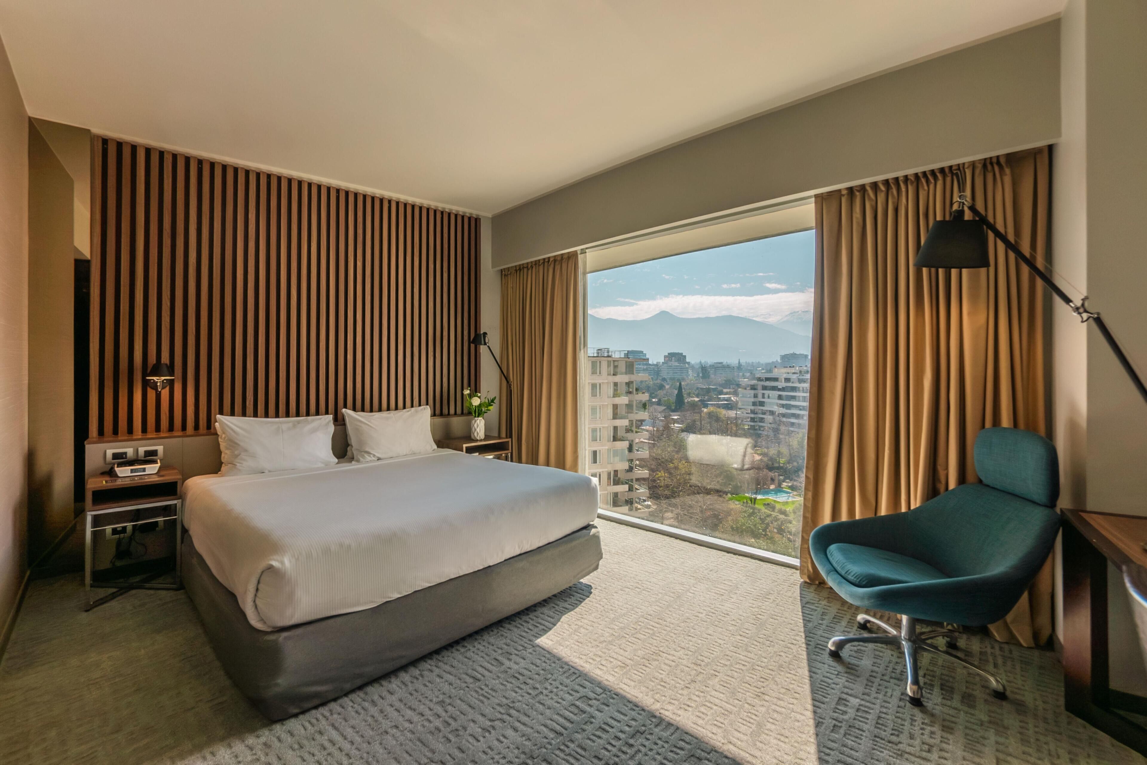 A hotel room with a bed, a desk, a chair, a large window with curtains, and a view of a cityscape.