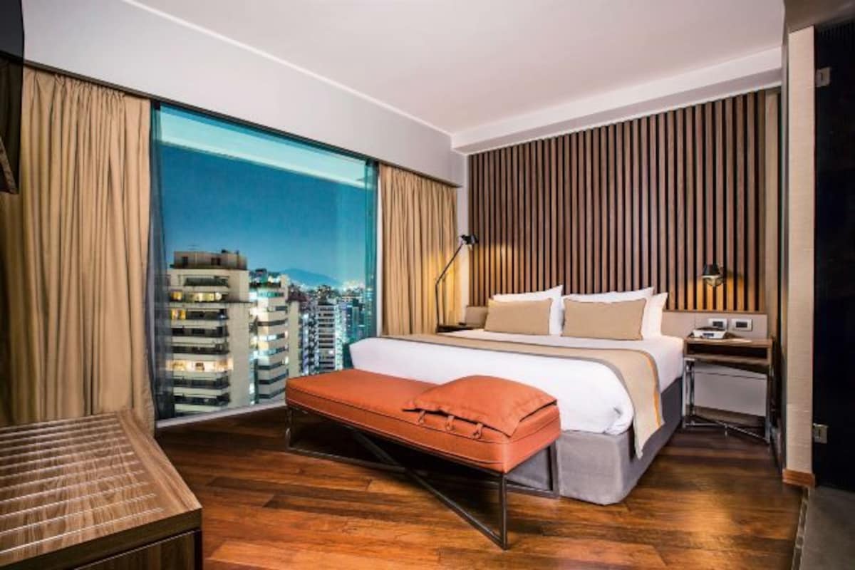 Una habitación de hotel moderna con una cama grande, un banco de madera, un escritorio y vistas de la ciudad a través de ventanas de gran altura.