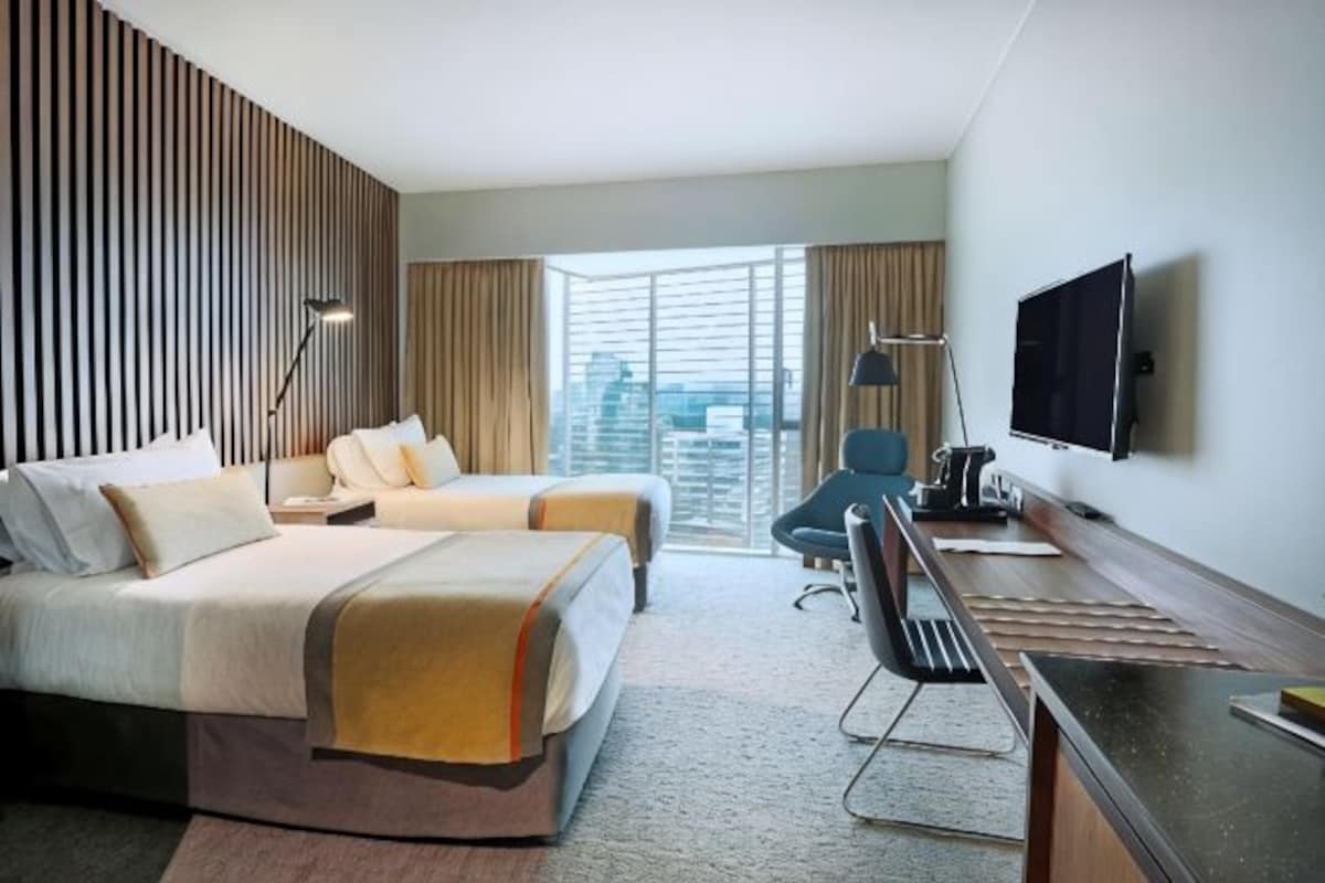 Habitación de hotel con cama, escritorio, silla, televisión y un amplio ventanal con vista a la ciudad.