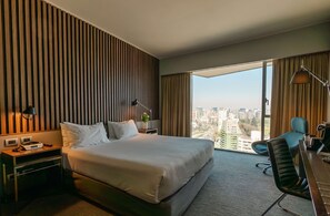 Habitación de hotel con una cama grande, un escritorio con silla, una lámpara y vistas a la ciudad.
