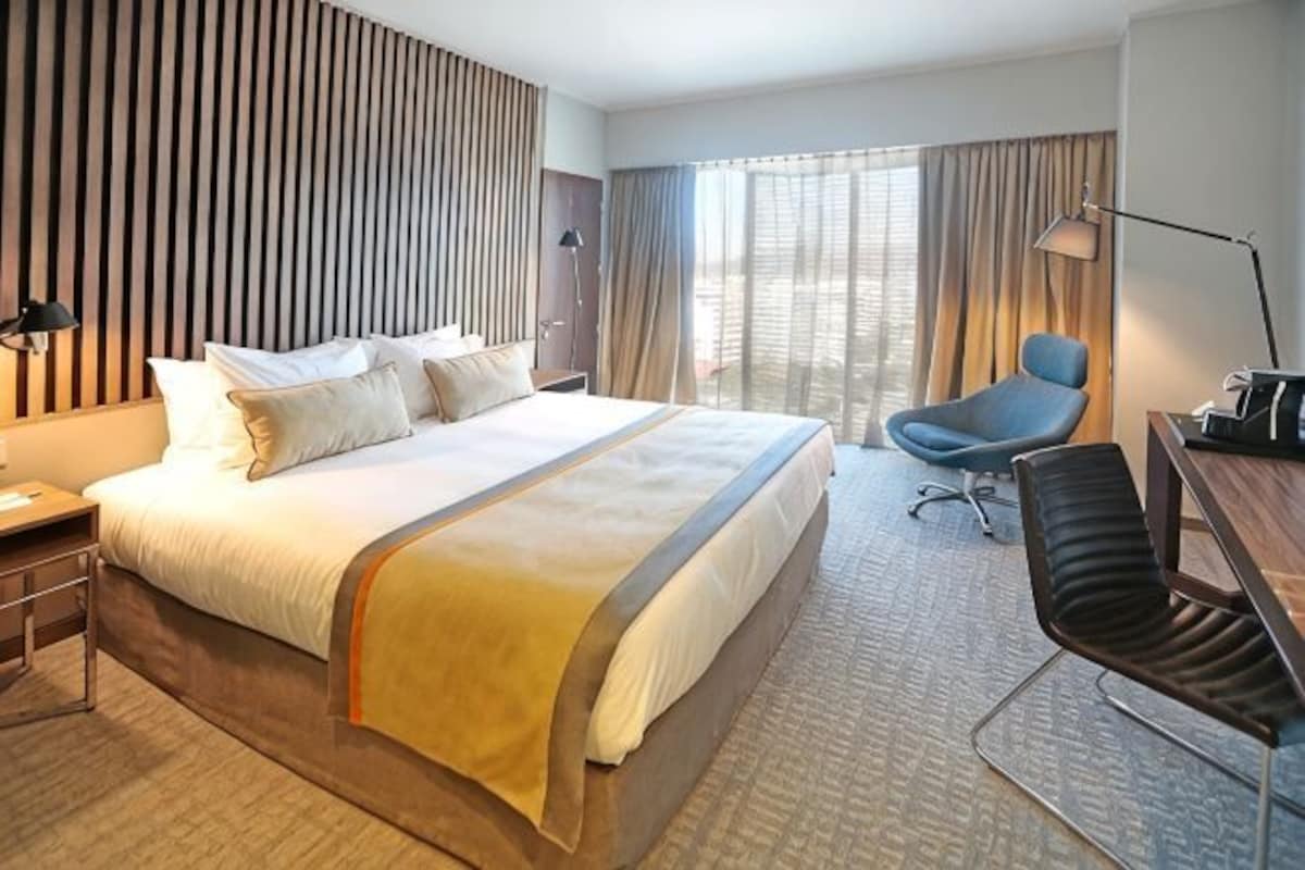 Una habitación de hotel con una cama grande, un escritorio con una silla, una lámpara y vistas al exterior.