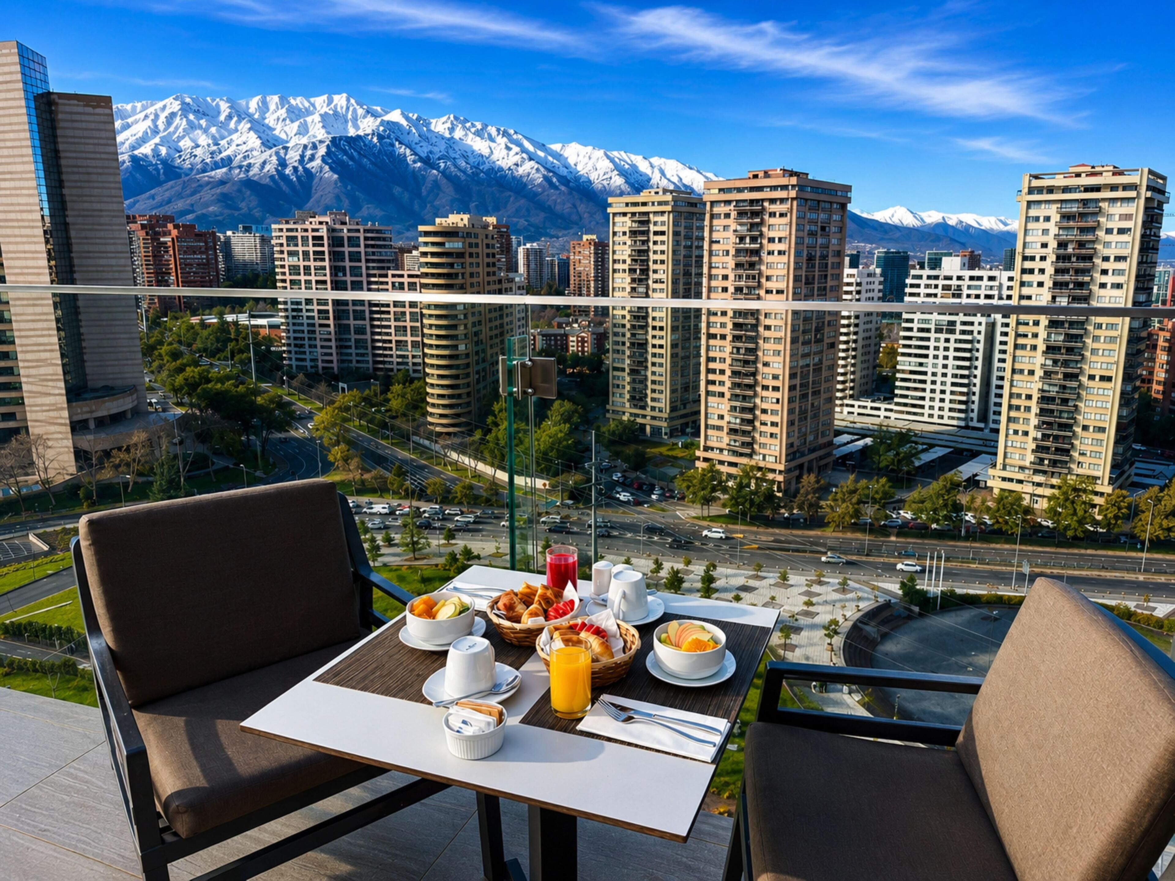 A balcony with a table set for breakfast, overlooking a cityscape with high-rise buildings and snow-capped mountains.