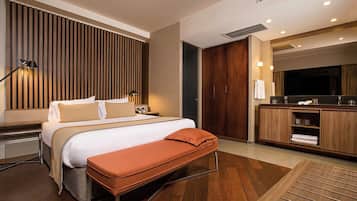 A modern hotel room with a large bed, a bench, a wooden wardrobe, a vanity with a mirror, and a television.