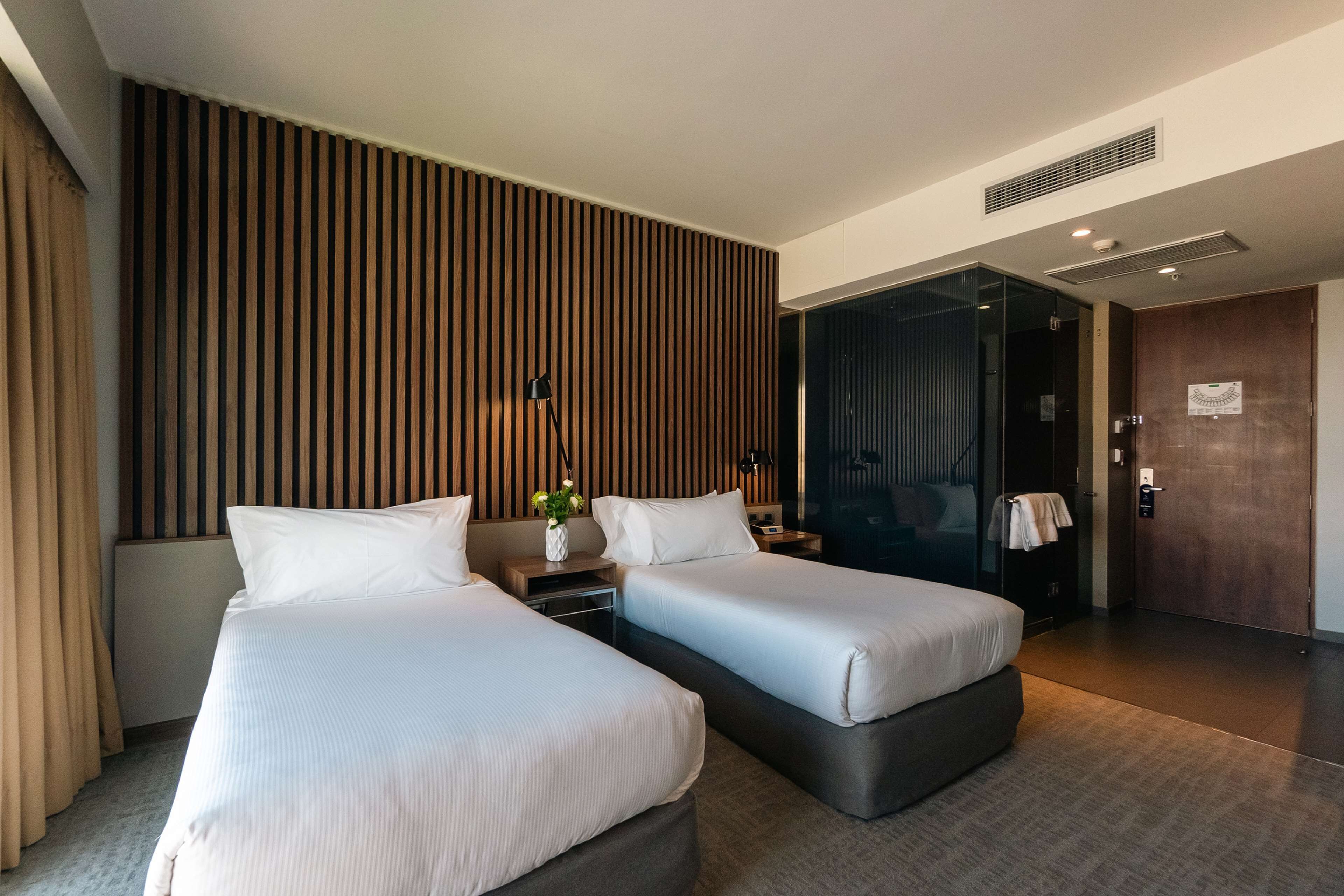 Una habitación de hotel moderna con dos camas, un muro de listones de madera, una mesita de noche con lámpara y una puerta que da a otra habitación.