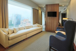 A modern hotel room with a sofa, a chair, a TV mounted on the wall, and a view of the city through large windows.